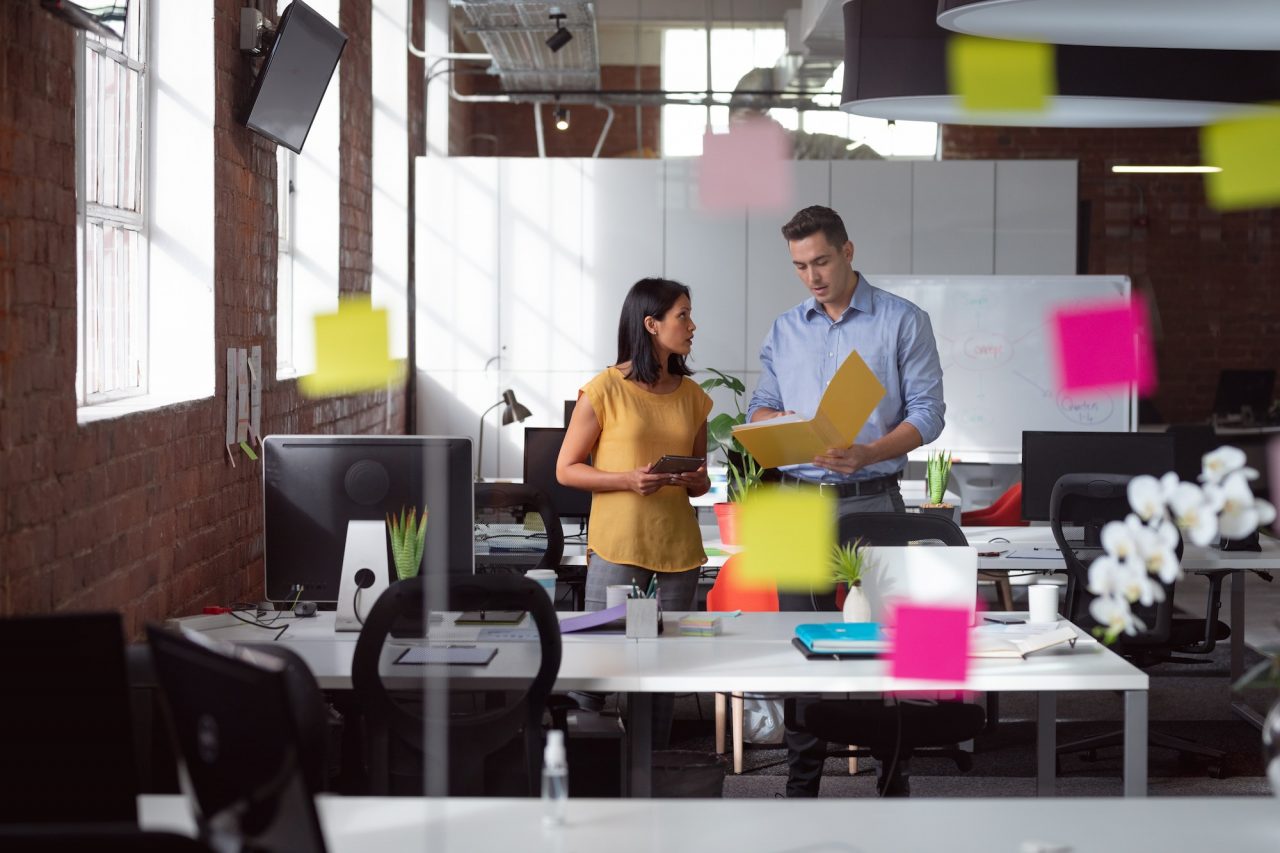 Caucasian male and female colleague discuss tablet and file, view through glass wall and memo notes